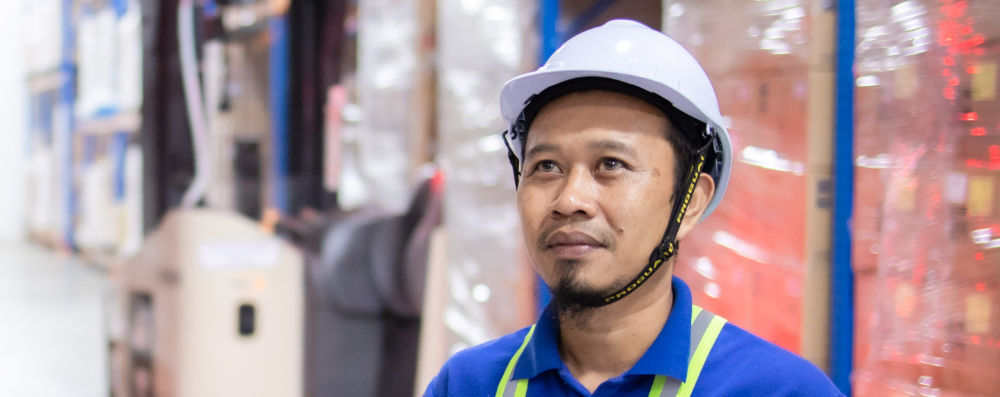 A warehouse worker, wearing a white hard hat, looking up, off-camera, with stacks of product behind him.